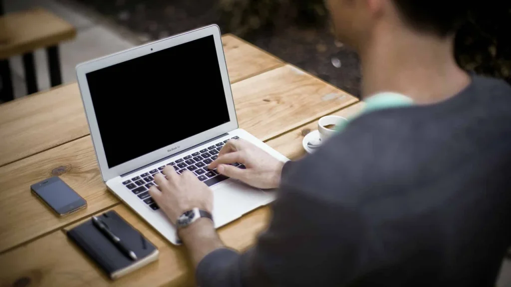 Munkatárs laptopon dolgozik egy asztalnál, mellette telefon és jegyzetfüzet.